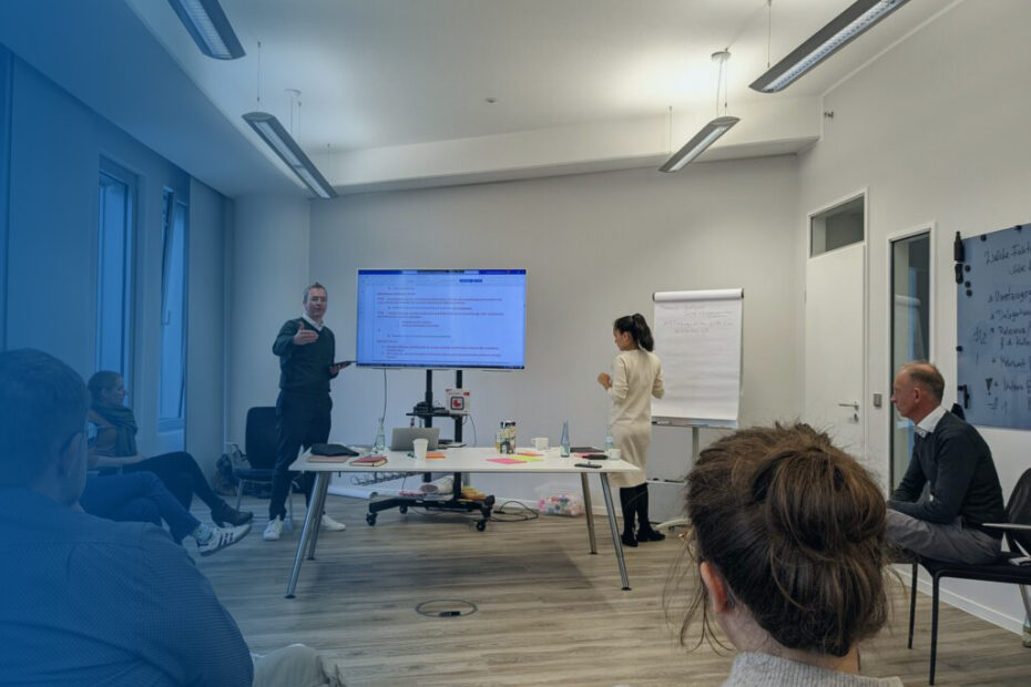 Ein Workshop zum Thema B Corp findet mit mehreren Menschen in einem Raum statt, die eine Präsentation auf einem Bildschirm sehen, während ein Redner daneben steht und erklärt.